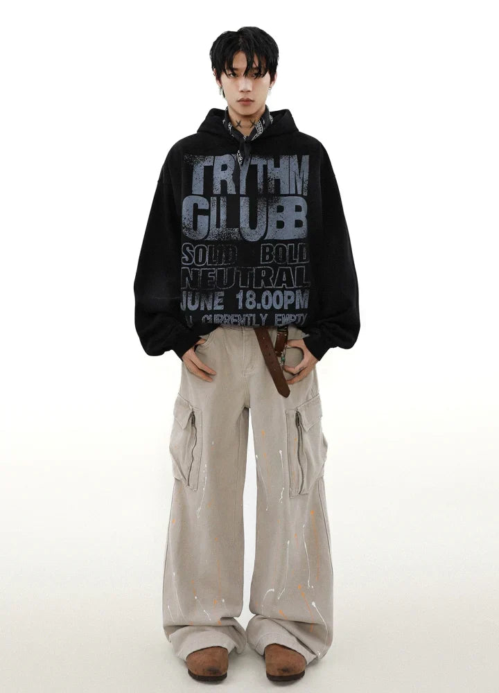 ベージュのカーゴパンツ、茶色の靴、そして太字で「RHYTHM CLUB、SOLID BOLD NEUTRAL、6月18日午後、現在空いています」と書かれた黒のキャンバス フーディを着て、白い背景に立っている人物。.