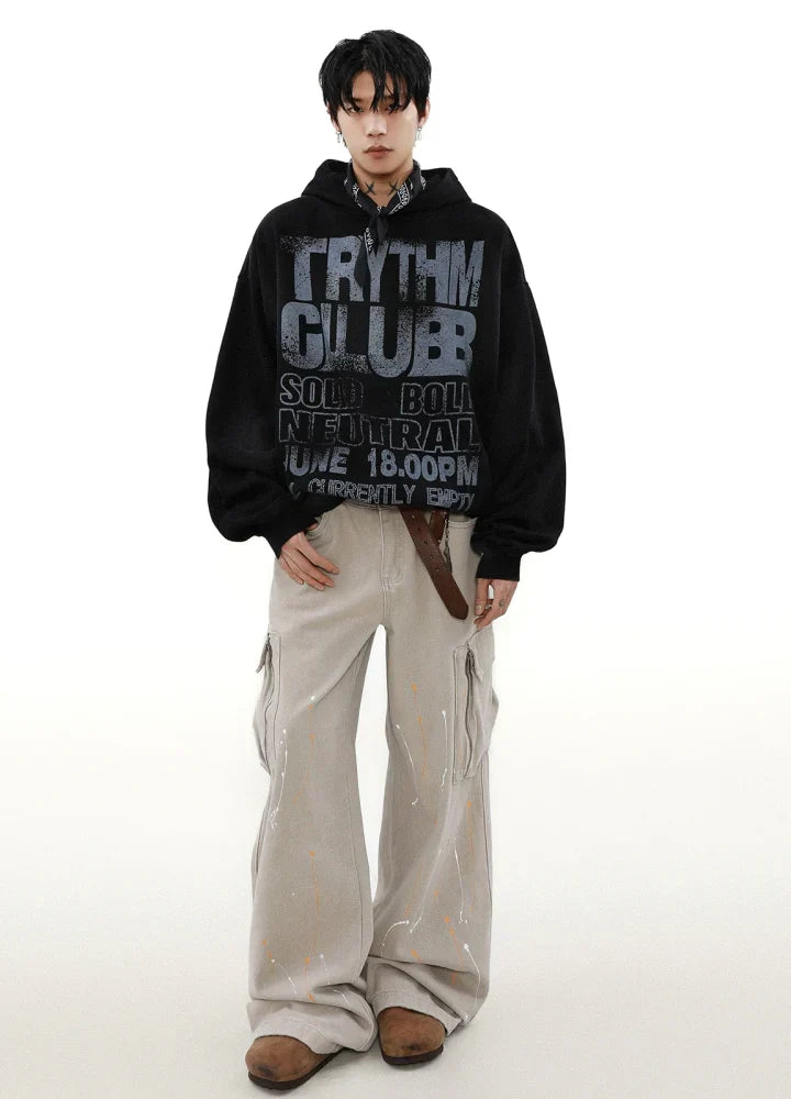 キャンバス フーディー、ベージュのワイドレッグ カーゴ パンツ、茶色の靴を履き、下にチェック柄のシャツを着た人物が、無地の白い背景に立っています。.